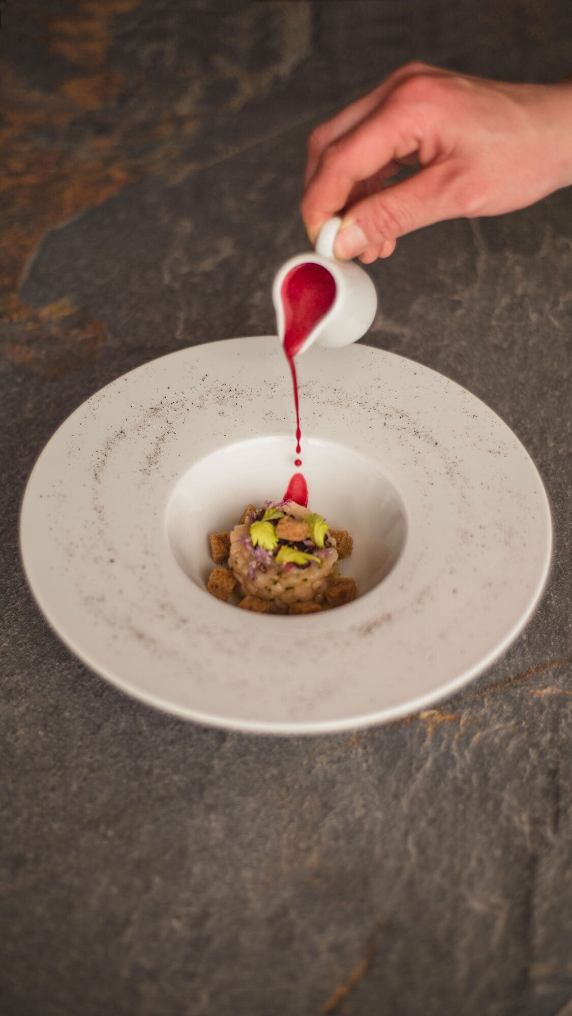 A person pouring a red sauce into a white bowl with a central dish.
