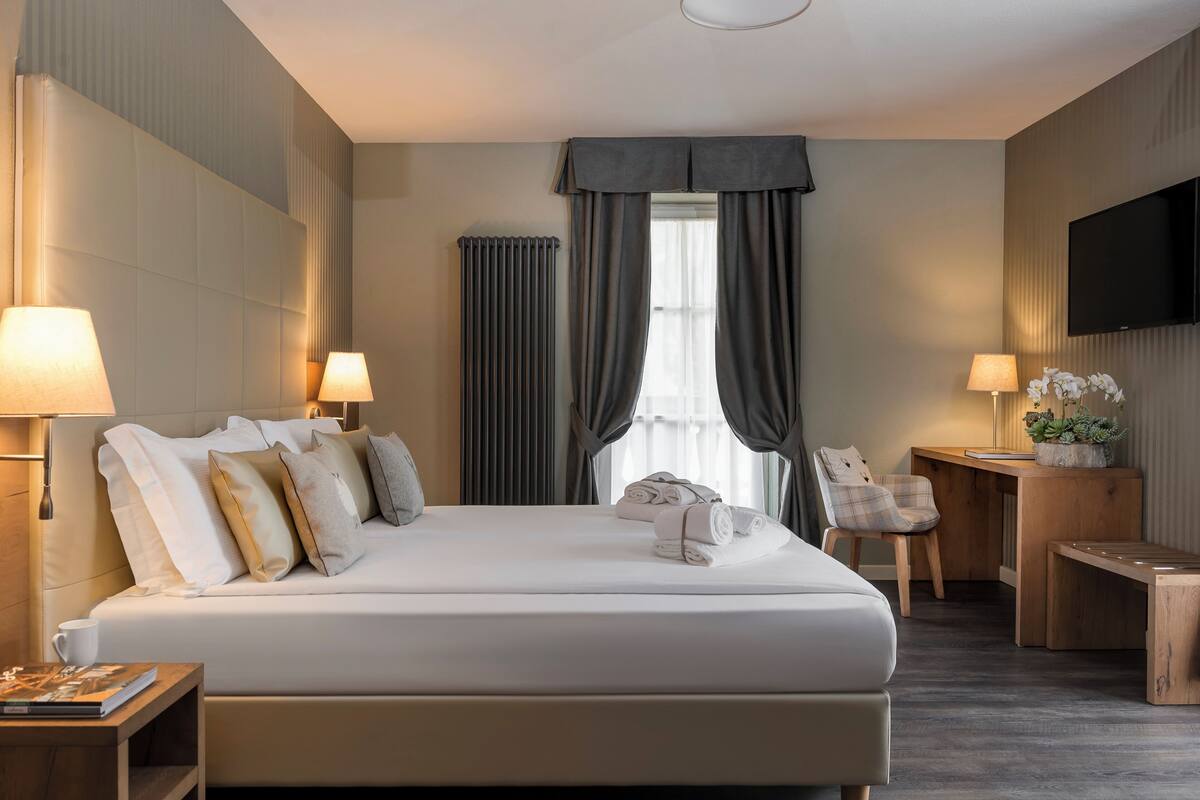 A hotel room with a large bed, two bedside lamps, a desk with a chair, a television, and a window with curtains.