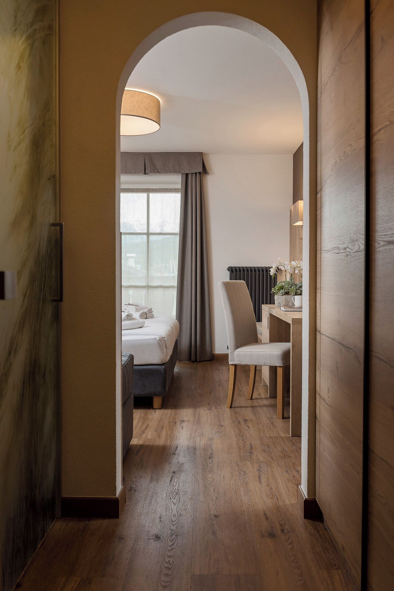 Habitación de hotel con piso de madera, una cama con cabecera gris, un escritorio con silla y un ventanal con cortinas.