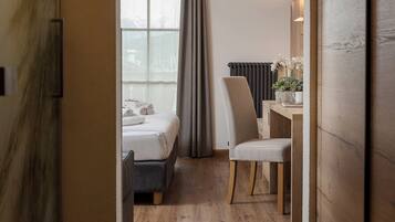 Habitación de hotel con piso de madera, una cama con cabecera gris, un escritorio con silla y un ventanal con cortinas.