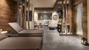 Une salle de spa moderne avec un sol en pierre, des murs en bois et un grand miroir. On y trouve deux fauteuils et une petite table sur laquelle est posée une plante.