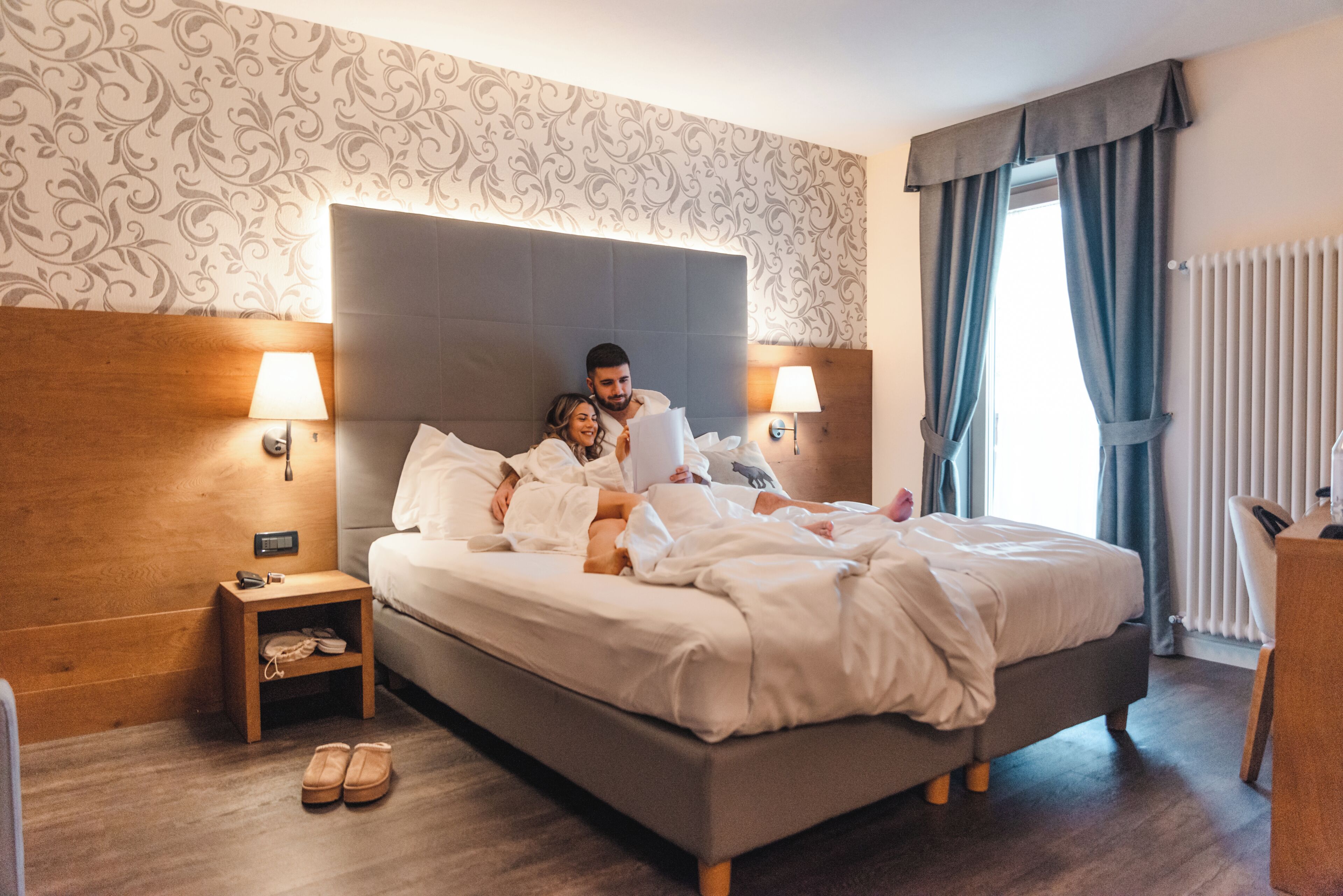 Una habitación de hotel con una cama grande, lámparas de noche, una mesita de madera y un par de pantuflas en el suelo.