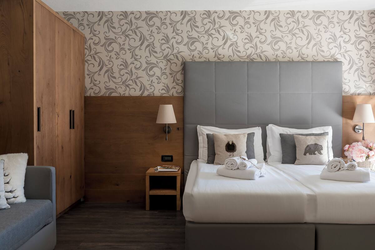 A hotel room with a large wardrobe, a bed with grey headboard, and patterned wallpaper.