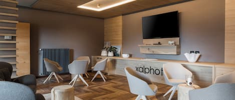 A modern interior with a wooden floor, grey and white chairs, a flat-screen TV mounted on the wall, and a reception desk with a sign reading "xalphot".