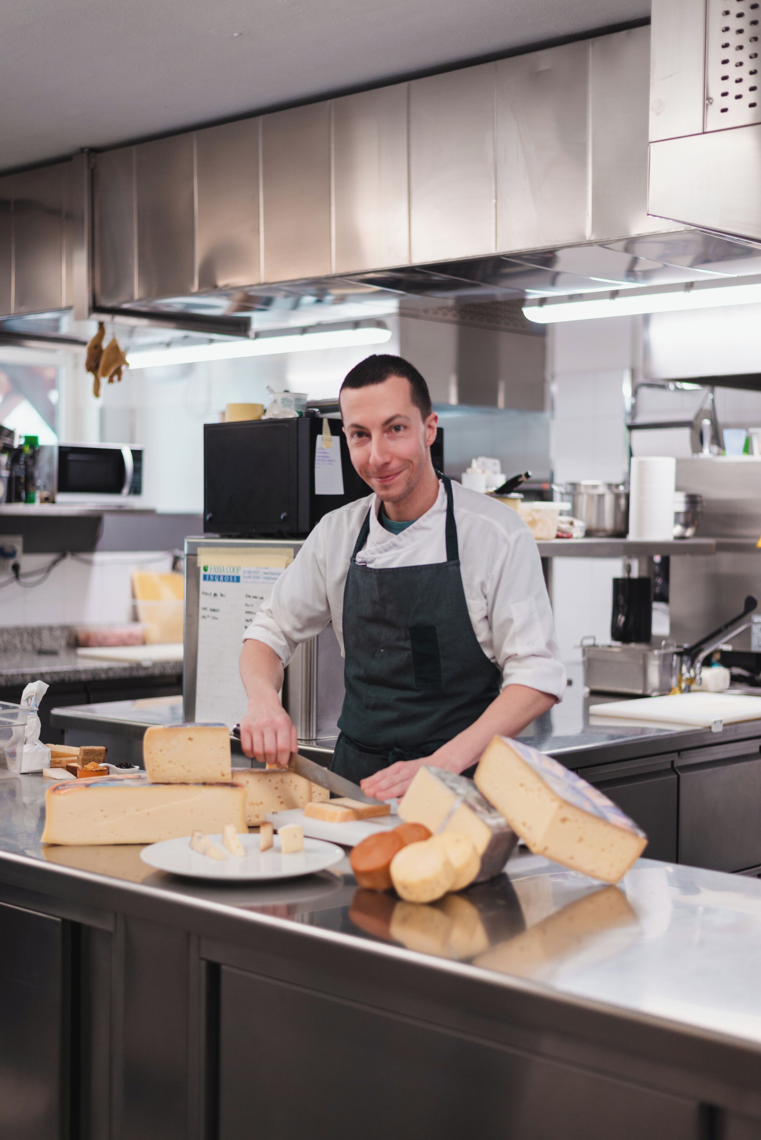 A chef in a professional kitchen with stainless steel appliances and a variety of cheeses and bread on the counter.