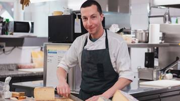 A chef in a professional kitchen with stainless steel appliances and a variety of cheeses and bread on the counter.