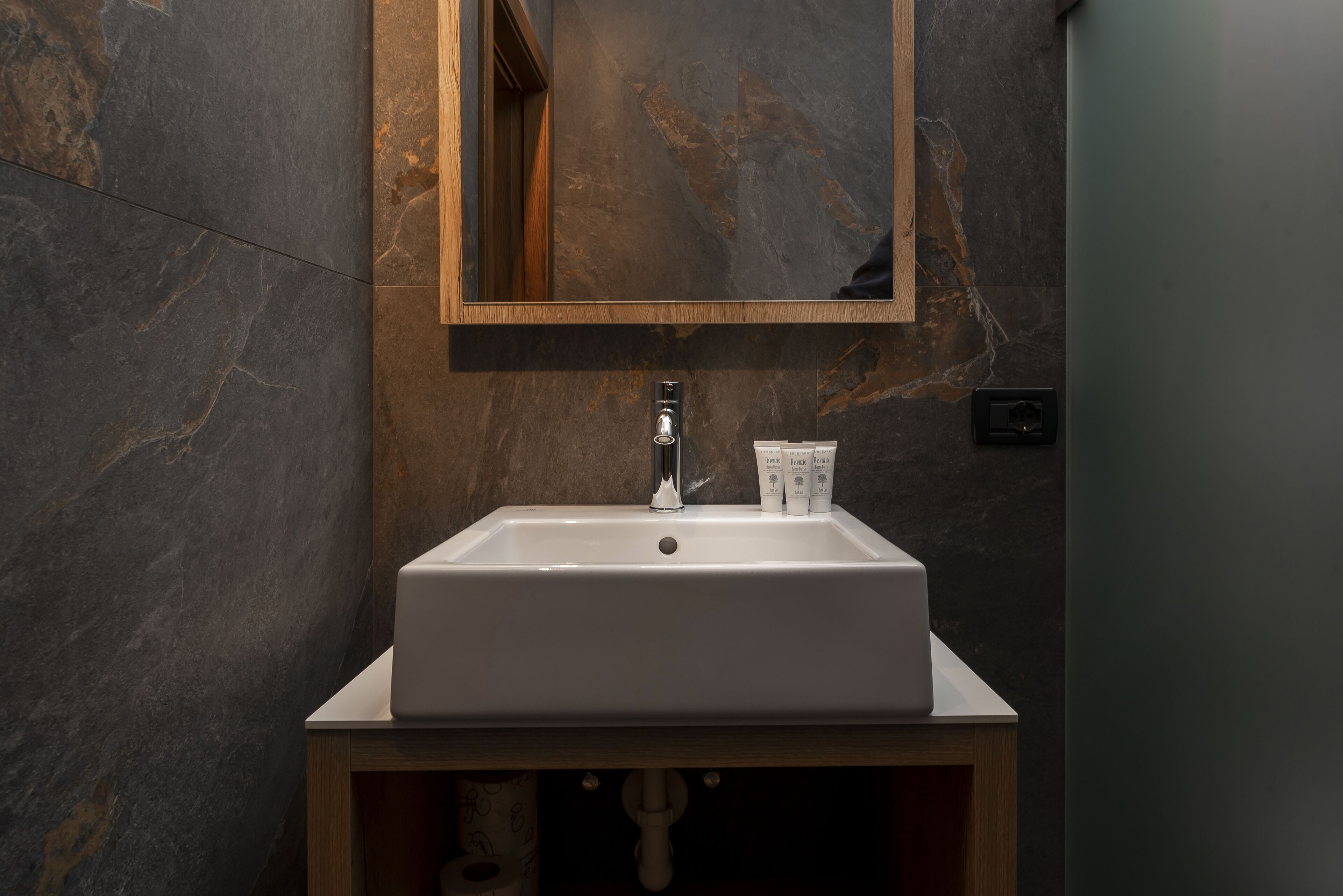 A bathroom with a white sink, a wooden vanity, a marble wall, and a mirror.
