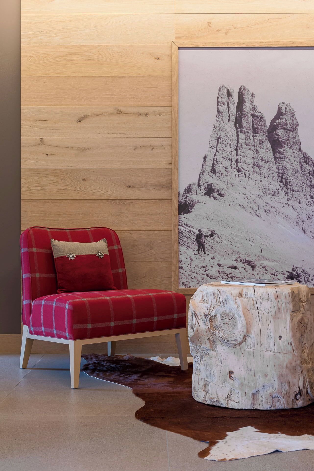 Una habitación con pared de madera, un sillón rojo a cuadros, un taburete de madera y un cuadro grande con un paisaje de montaña.