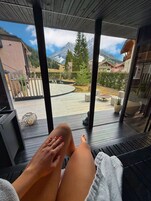 A person relaxing on a deck with a view of a mountainous landscape, a pool, and a fenced garden area.