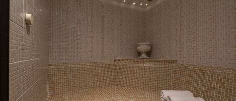A corner bathtub with mosaic tile walls and a white towel on the seat.