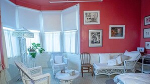 Una habitación con mobiliario blanco de mimbre, un sofá blanco y una cama. Hay cuadros en la pared y una planta en maceta.