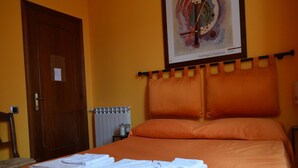 Een hotelkamer met een bed, oranje beddengoed, een radiator, een deur en een lijst met een kunstwerk aan de muur.