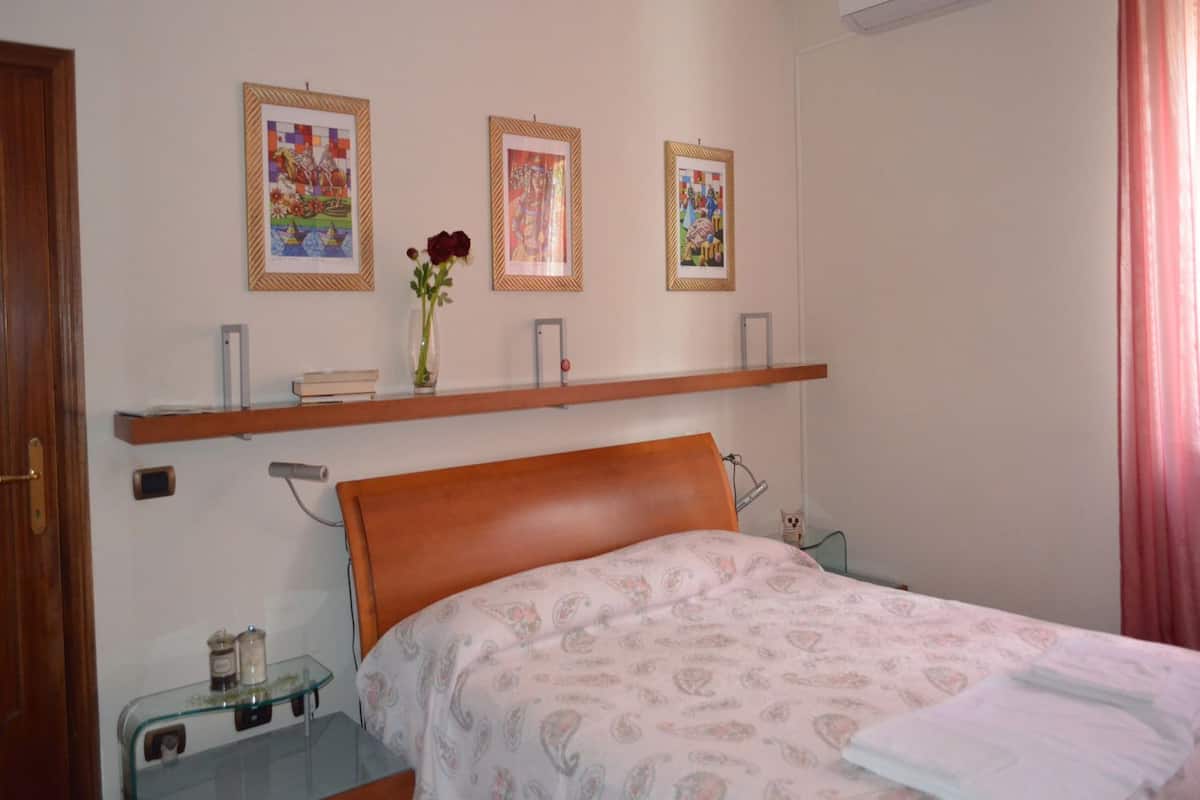 Un dormitorio con cama, mesita de noche, estante con marcos de fotos y un florero con flores.