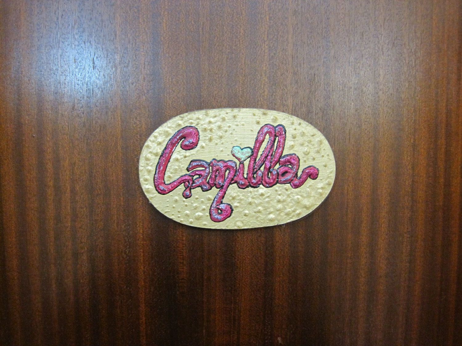 Een houten oppervlak met een ovaal sticker met reliëf waarop ‘Camilla’ in sierletters staat.