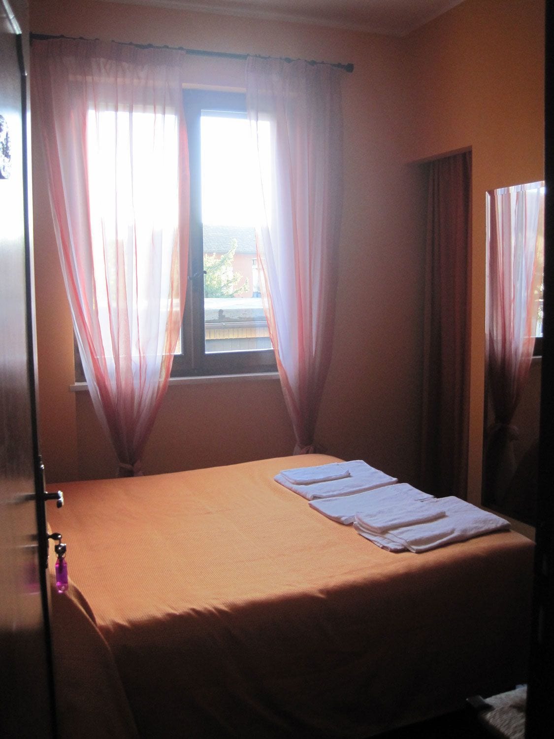 Una stanza con un letto, una finestra con tende leggere e vista sugli edifici.