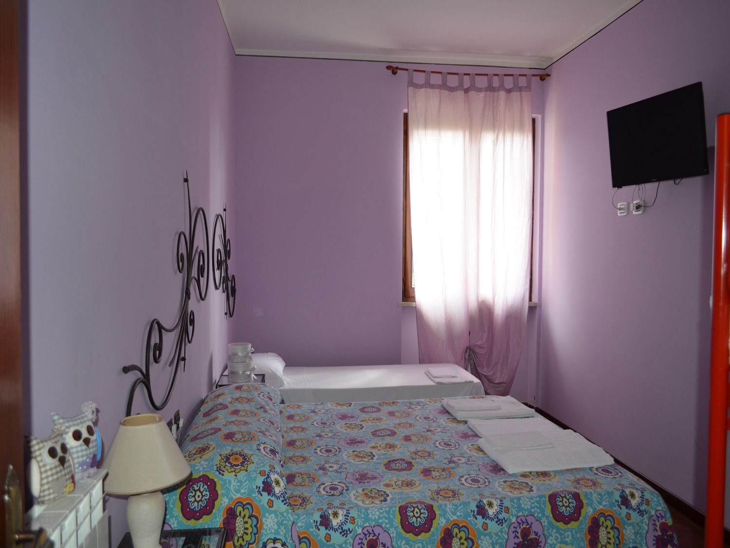 Quarto com duas camas, janela com cortinas leves, televisão de tela plana na parede e abajur em uma mesa de cabeceira.