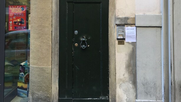 Une porte noire avec un heurtoir en métal, une plaque portant le numéro « 79 », et une petite fenêtre ornée d’une grille décorative.
