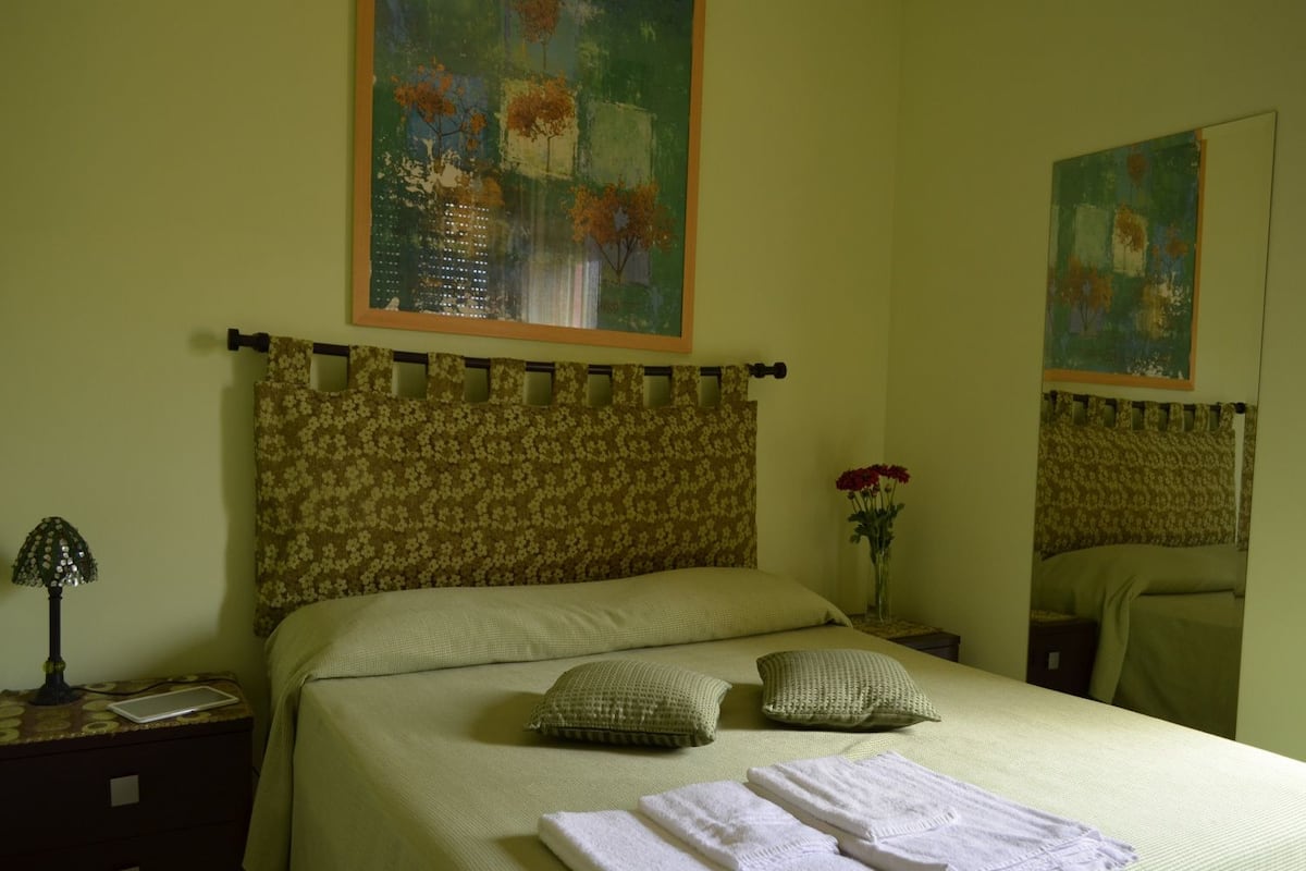 A neatly made bed with a floral headboard, a bedside table with a lamp, and a vase with flowers.