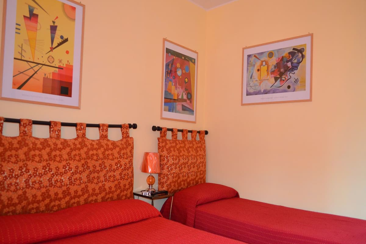 A room with two beds, each with red bedding, a small table with a lamp, and three abstract paintings on the walls.