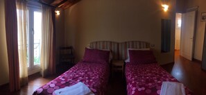 A room with two beds, each with a striped headboard and pink bedding. There is a window with curtains, a chair, and a door on the right side. - Amorino (Florence)