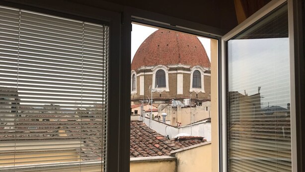 Une vue à travers une fenêtre munie de stores, dévoilant un bâtiment surmonté d’un dôme rouge et doté d’un toit en tuiles.