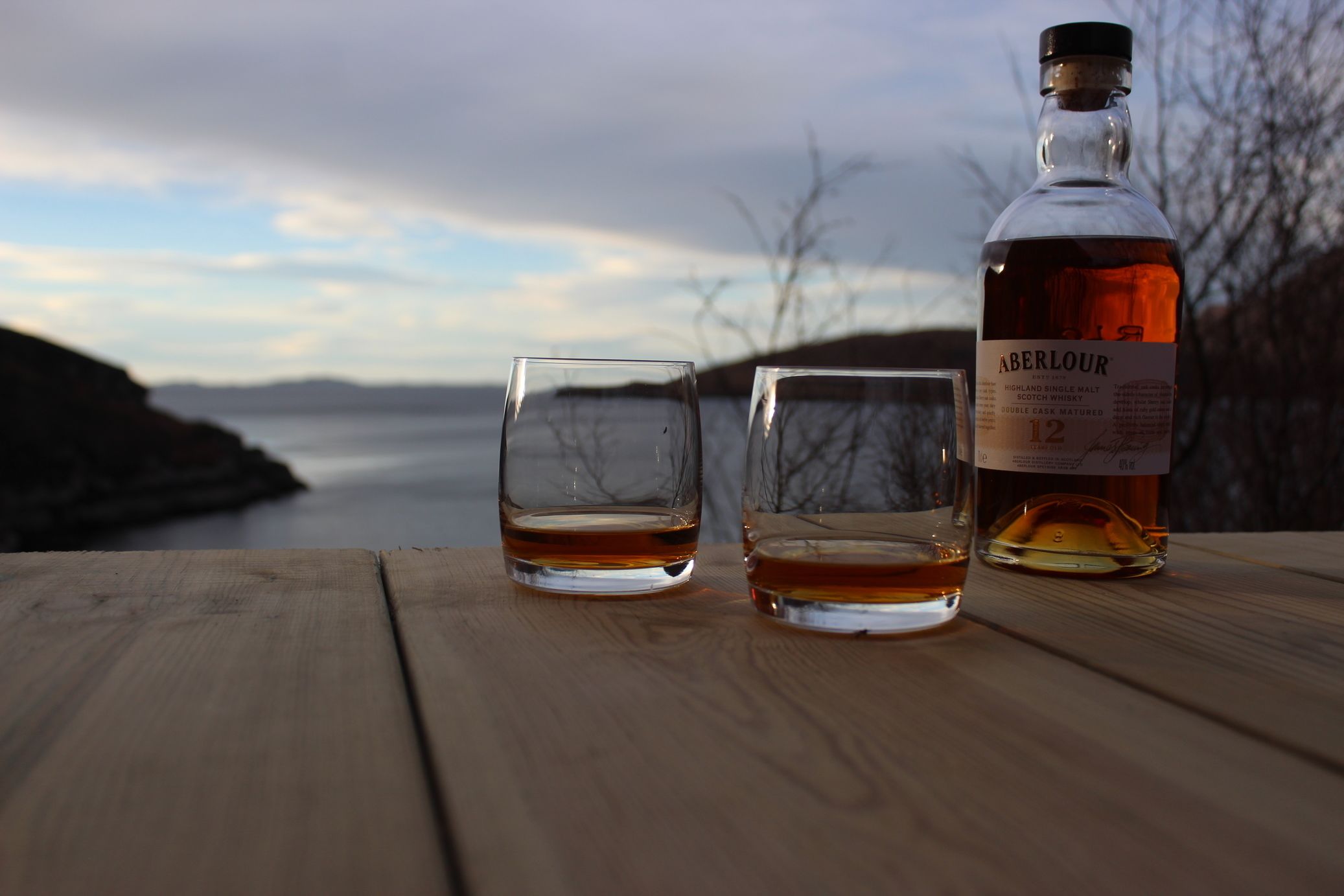 Two glasses of Aberlour whisky on a wooden table with a bottle beside them, overlooking a body of water and hills.