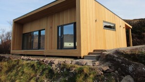 Une cabane moderne en bois, dotée de grandes portes-fenêtres coulissantes, d’une petite fenêtre et de fondations en pierre.