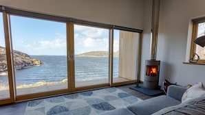 Une pièce chaleureuse avec une grande fenêtre donnant sur la mer, une cheminée moderne et un coin salon confortable.