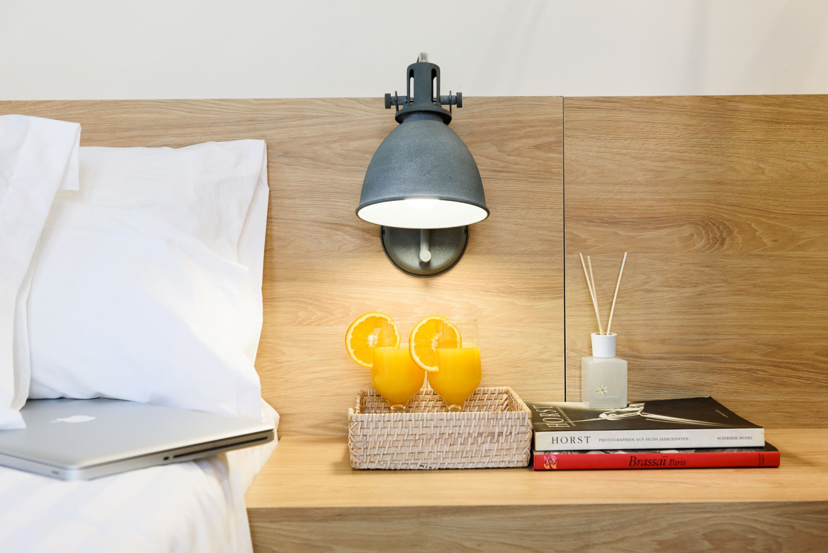 A neatly made bed with white linens, a laptop on the bed, a wall-mounted lamp, a tray with two glasses of orange juice, a book titled 'Brassai Paris', and a reed diffuser on a wooden nightstand.