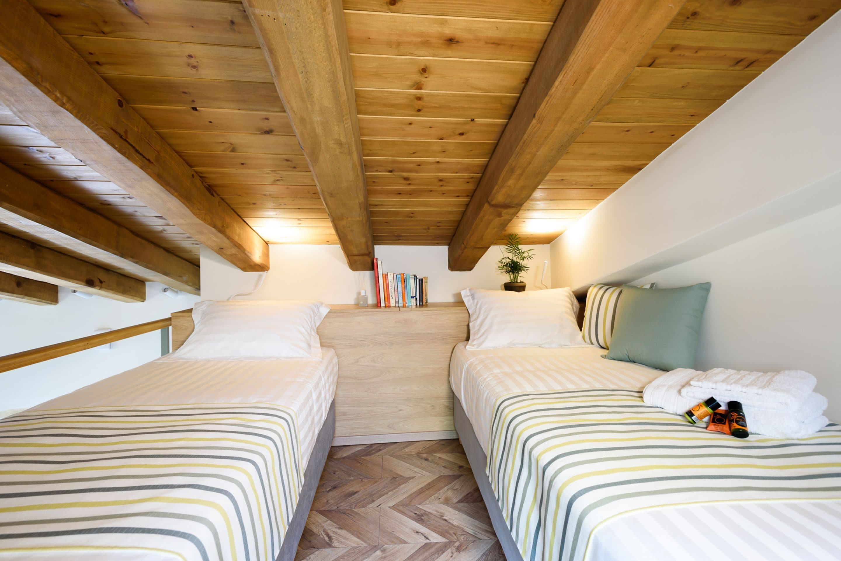 A room with two beds, wooden beams on the ceiling, a herringbone floor pattern, and a small plant on the wall.