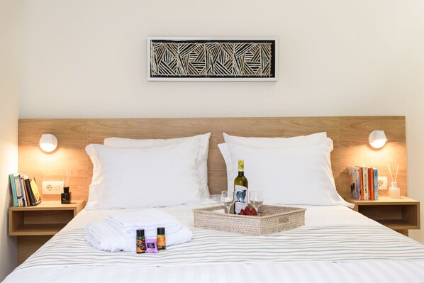 Una cama bien hecha con sábanas blancas, una botella de vino, copas y una cesta de fruta sobre la mesita de noche.