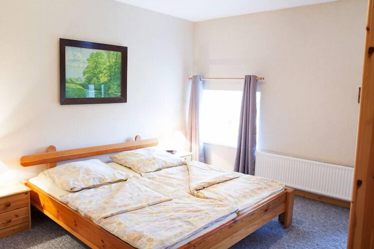 Un dormitorio con cama de madera, una mesita de noche, un cuadro enmarcado en la pared y una ventana con cortinas.
