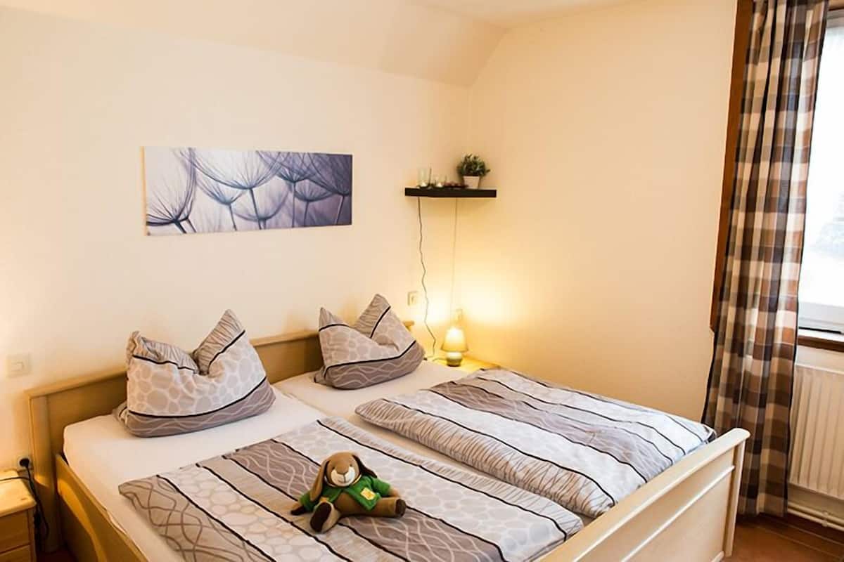 Una cama bien hecha con ropa de cama estampada, una estantería de pared con objetos decorativos y una ventana con cortinas.
