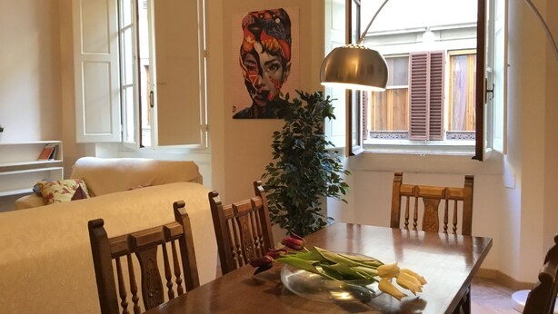 A dining area with a wooden table, chairs, a vase with flowers, a lamp, and a window with shutters. - Battistero (Florence)
