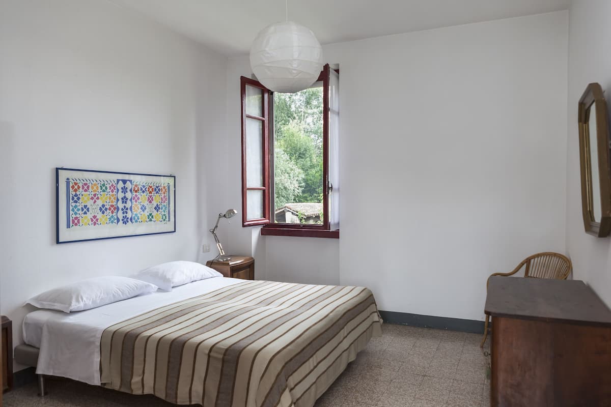 Un dormitorio con cama, escritorio, silla, ventana con persiana abierta y lámpara colgante.