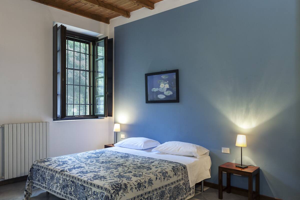 Un dormitorio con cama, dos mesitas de noche con lámparas, ventana con marcos negros, radiador y un cuadro en la pared.