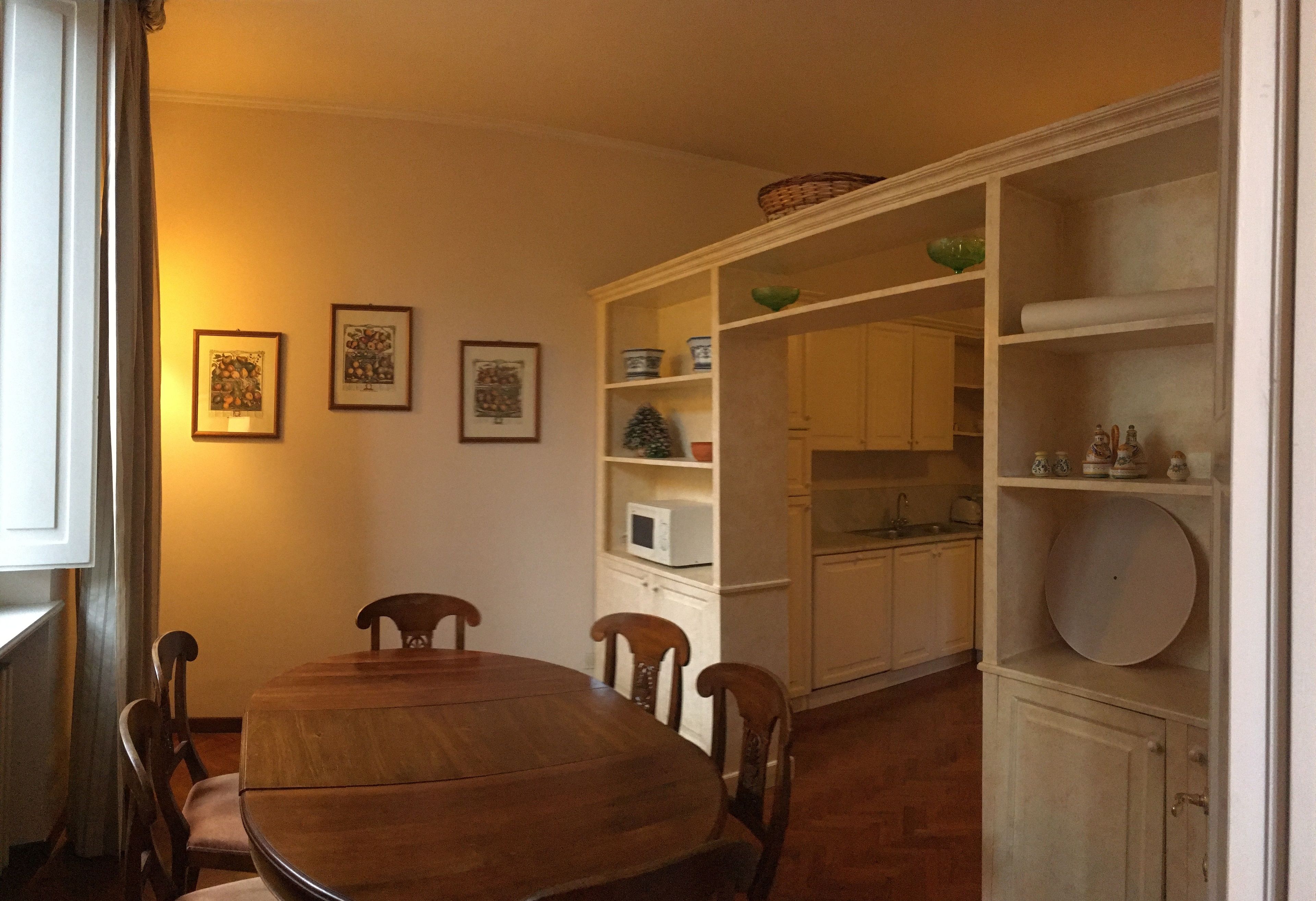 A dining area with a wooden table and chairs, a white microwave, and a shelf with decorative items.