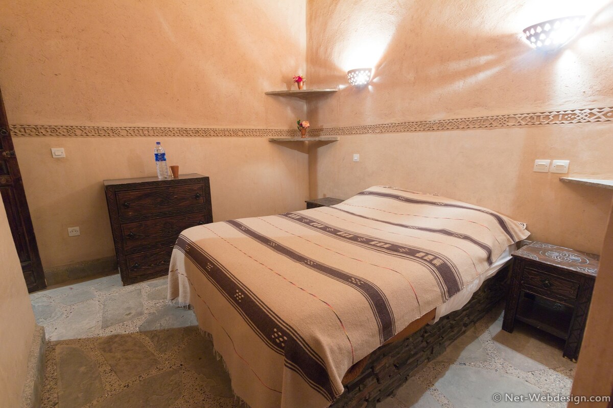 Un dormitorio con cama, cómoda, mesita de noche, estante con un florero y una botella de agua.
