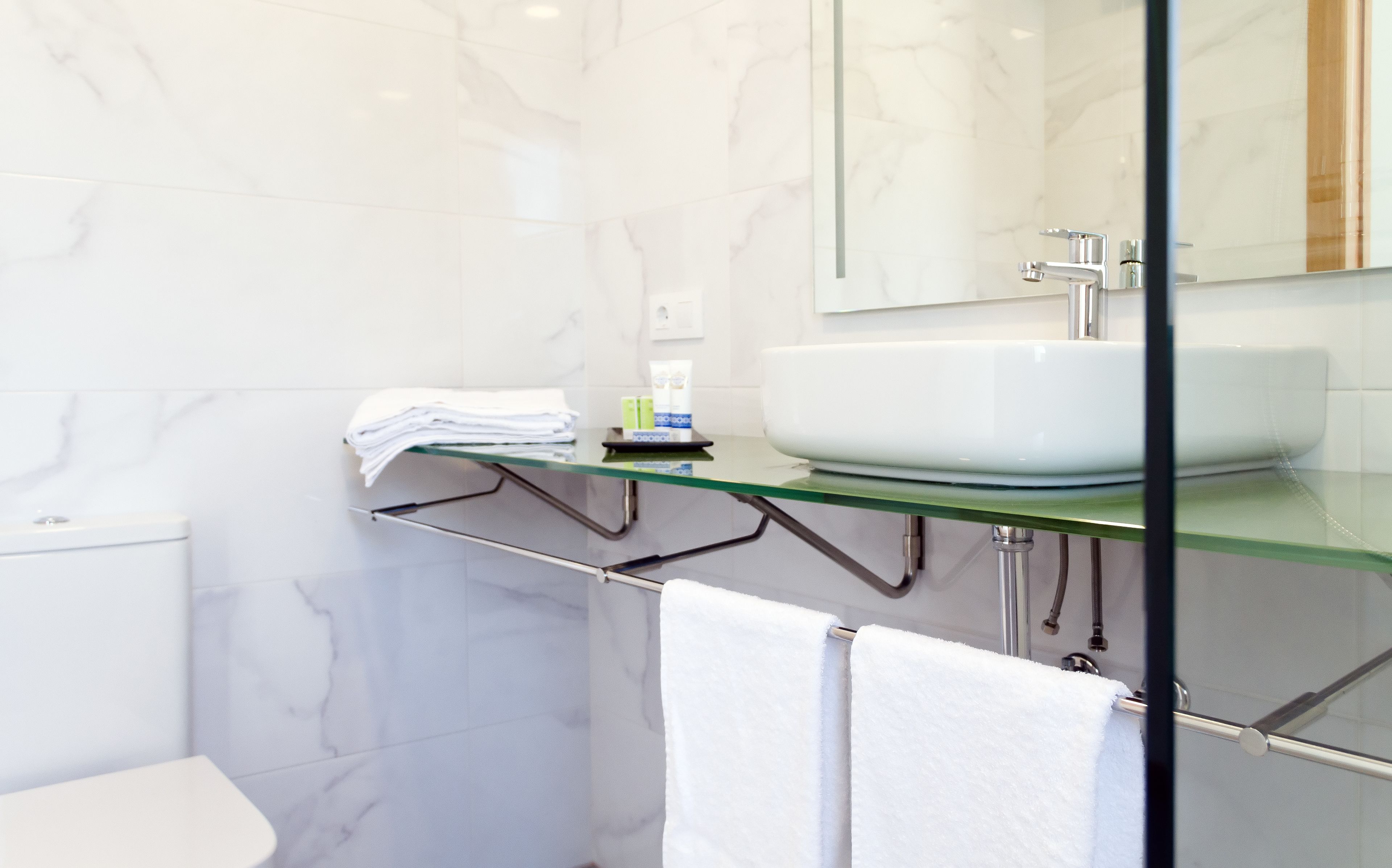 Un baño moderno con repisas de vidrio que sostienen artículos de tocador, un lavamanos blanco y un inodoro.