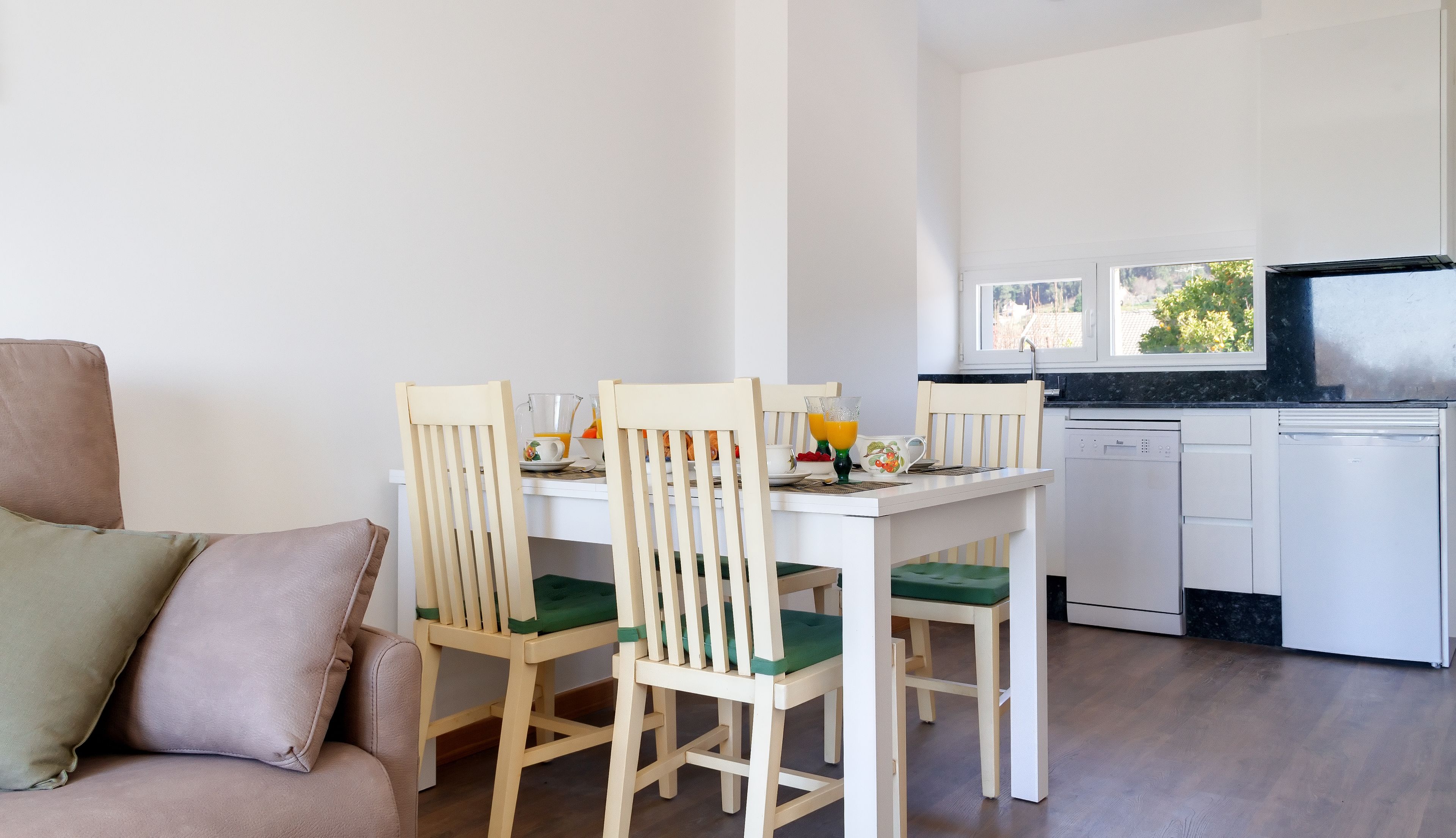 A dining area with a white table and chairs, a kitchen with white appliances, and a view of trees outside.