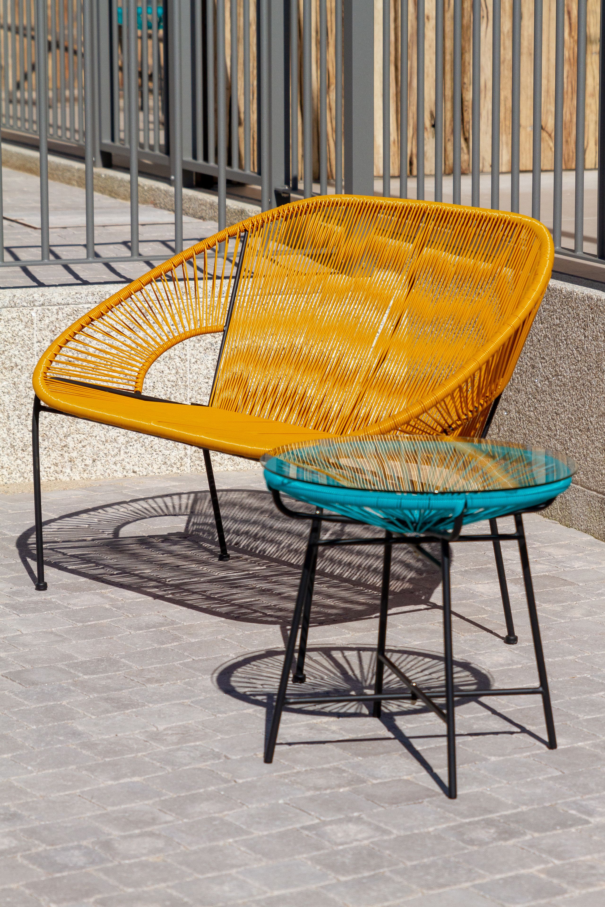 A yellow woven chair and a turquoise woven table on a paved surface with a metal fence in the background.