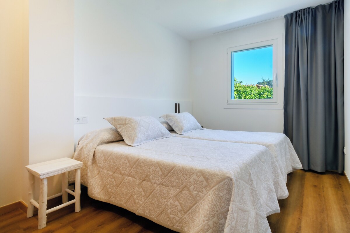 Un dormitorio con una cama, una mesita de noche pequeña y blanca, una ventana con vistas a zonas verdes y una cortina gris.