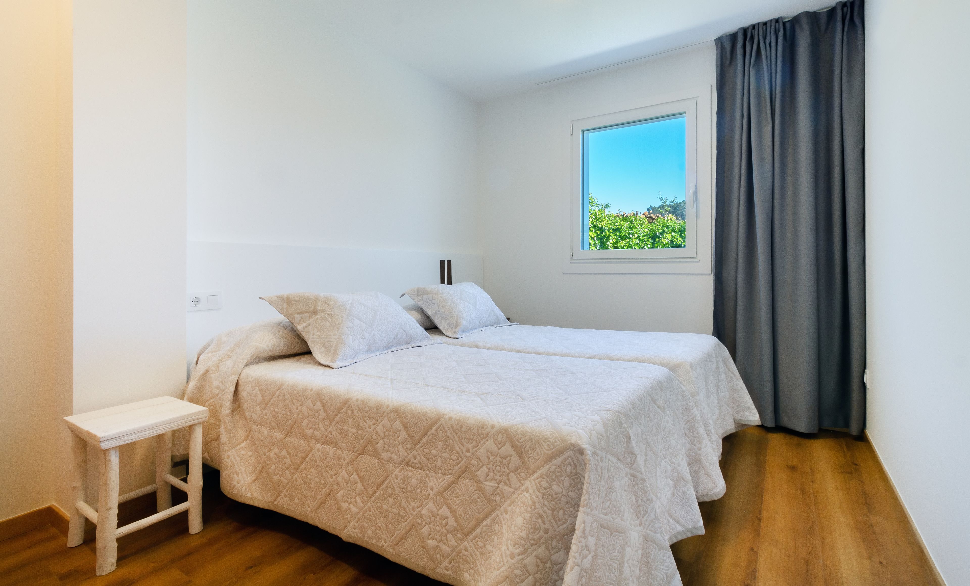 A bedroom with a bed, a small white nightstand, a window with a view of greenery, and a grey curtain.