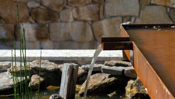 Una estructura de madera con agua que fluye de un caño hacia un estanque rodeado de rocas y un muro de piedra.