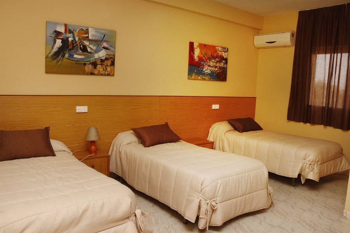 Quarto de hotel com três camas, cabeceira de madeira, duas pinturas nas paredes, ar condicionado e uma janela com cortinas.