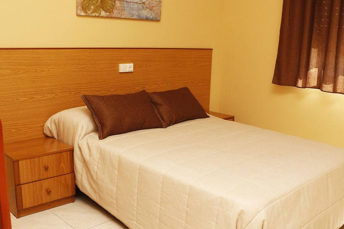 Cama bem arrumada com cabeceira de madeira, mesa de cabeceira com gaveta e interruptor de luz na parede.