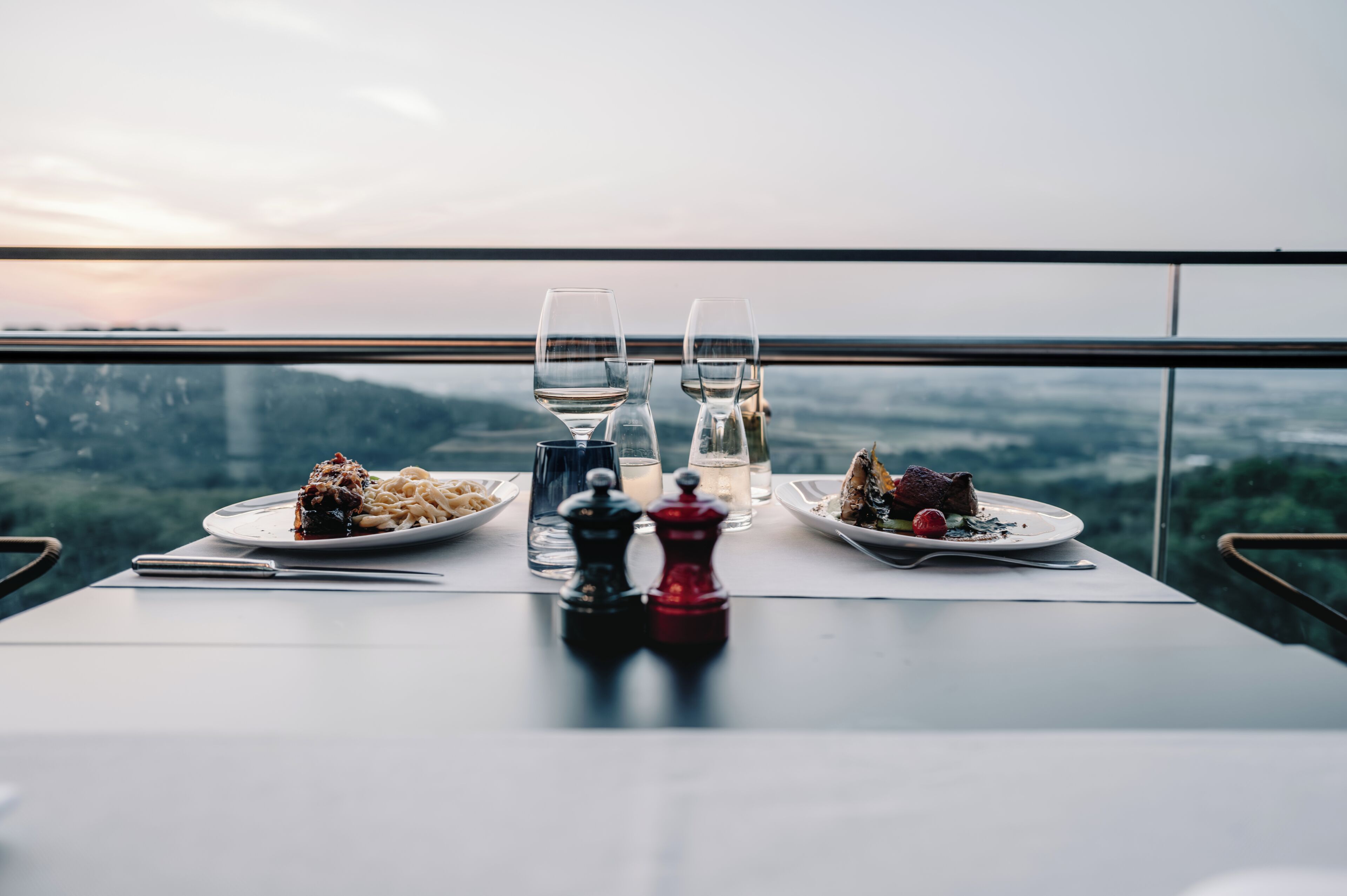 Una mesa de comedor con dos platos de comida, dos copas de vino y un juego de salero y pimentero en un balcón con vistas a un paisaje urbano.
