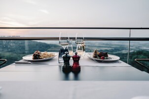 Una mesa de comedor con dos platos de comida, dos copas de vino y un juego de salero y pimentero en un balcón con vistas a un paisaje urbano.