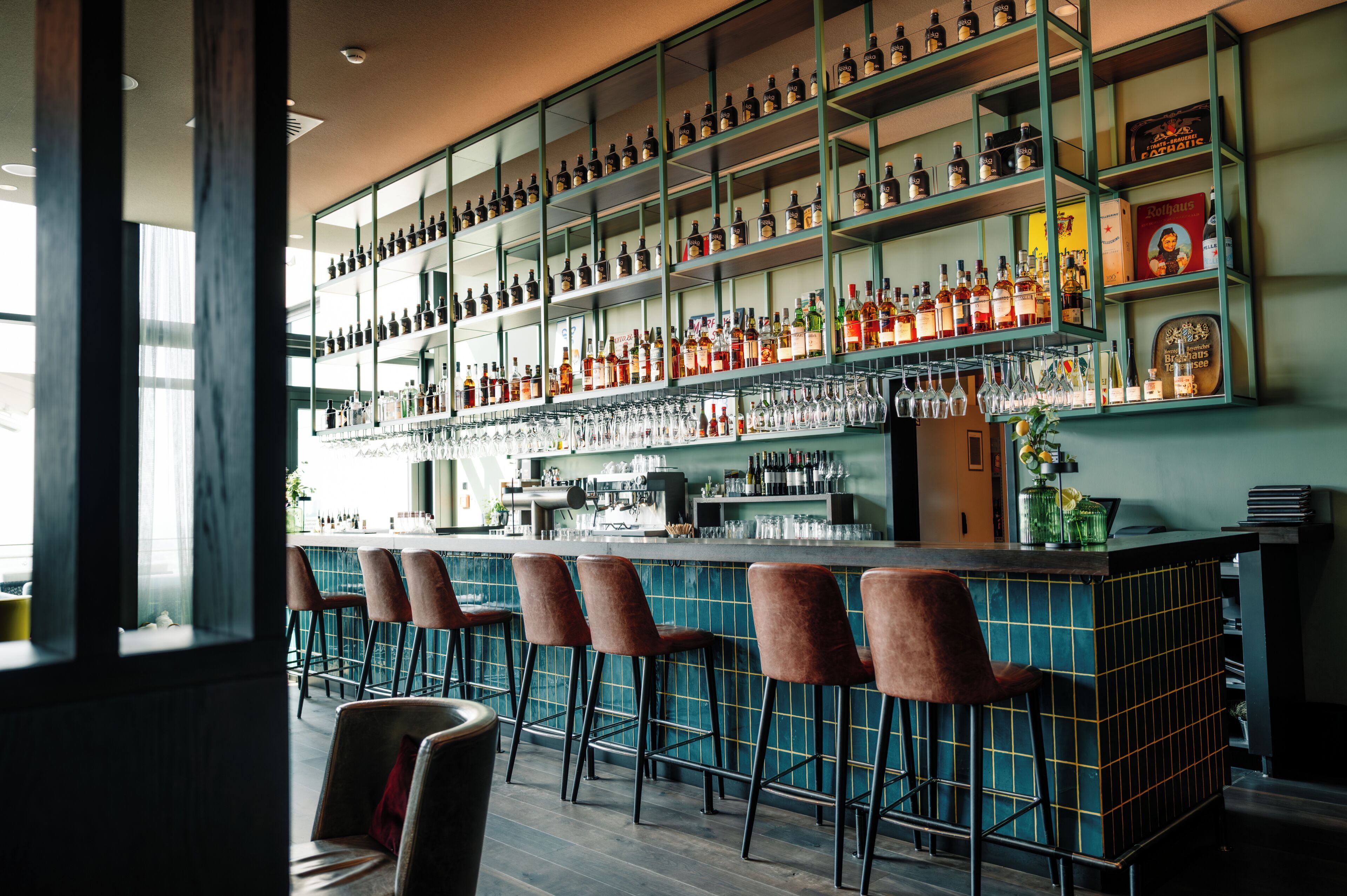 Una zona de bar con múltiples estantes llenos de botellas de licores, una barra y varios taburetes.