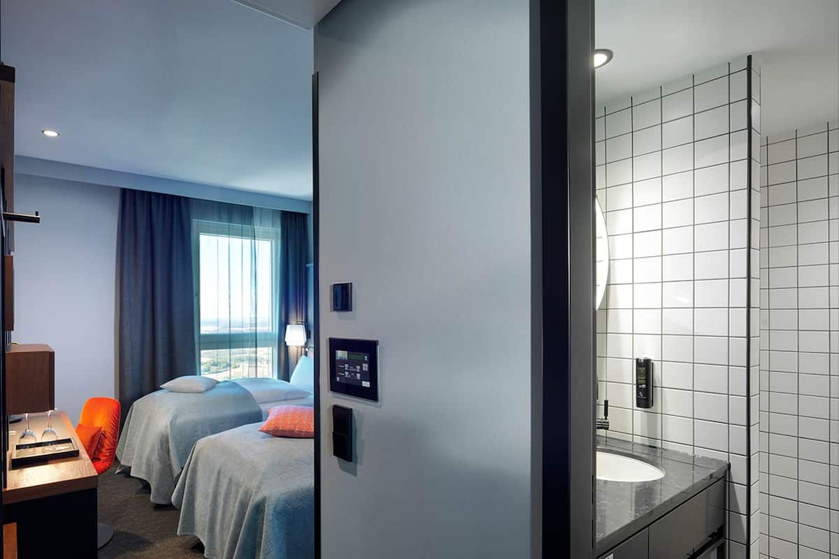 A hotel room with a bed, a desk, a chair, a window with curtains, a wall-mounted control panel, and a bathroom with a sink and tiled walls.